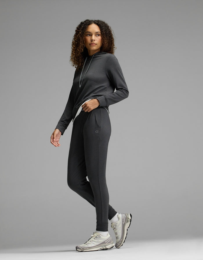 Wearing the Cozy Earth Women’s Brushed Bamboo Hoodie & Jogger Set in dark gray, a woman with curly hair pairs her outfit with white sneakers, standing against a plain gray background and gazing slightly to the side.|Color:Charcoal