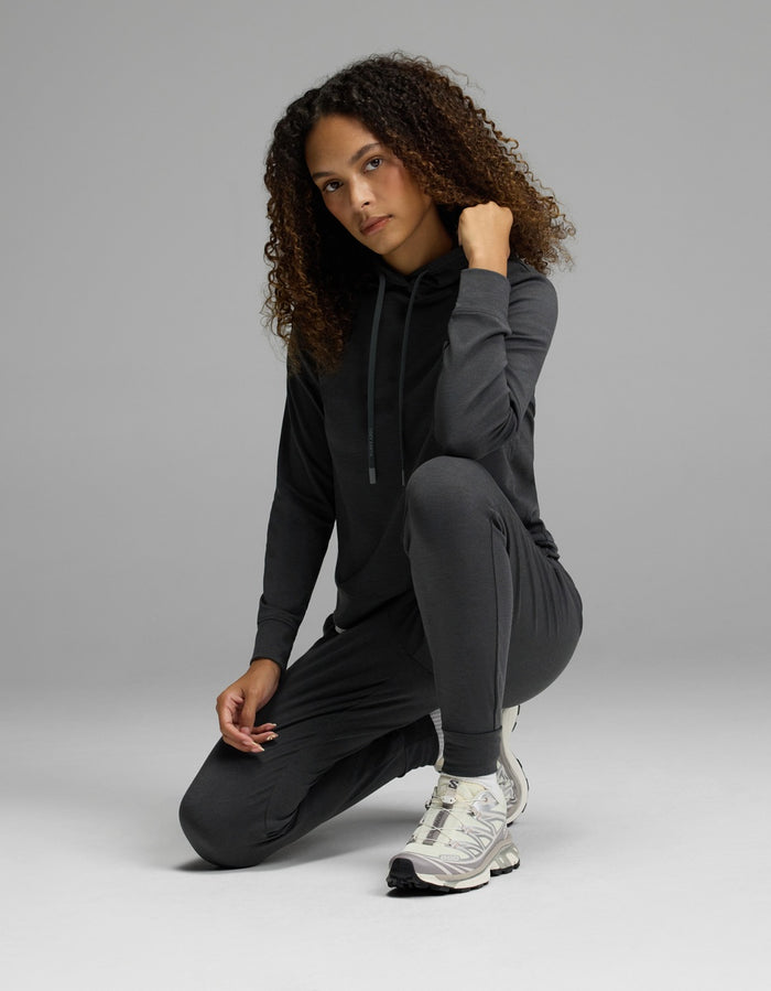 A woman with curly hair kneels on one knee against a plain gray background, wearing the Cozy Earth Women’s Brushed Bamboo Hoodie & Jogger Set in a dark color. She rests one hand on her chin and looks at the camera, completing her look with white sneakers.|Color:Charcoal