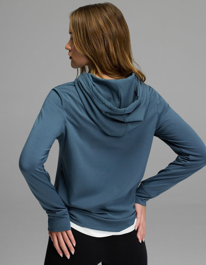 A person with long, light brown hair is seen from behind, wearing the Cozy Earth Women’s Brushed Bamboo Hoodie in blue and black pants, with one hand on their hip.|Color:Dawn