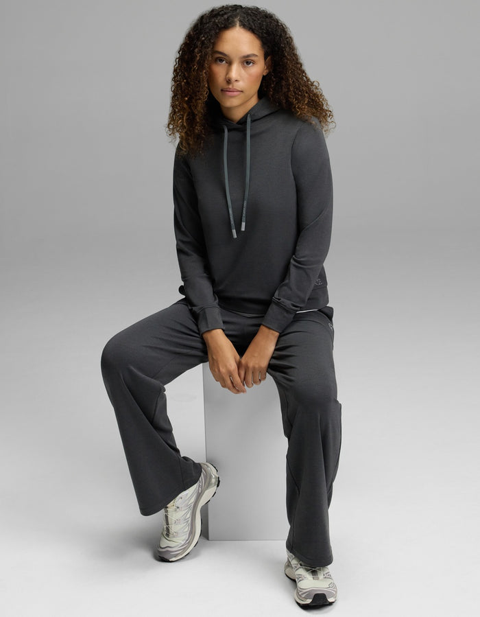 A woman with curly hair wears the Cozy Earth Women’s Brushed Bamboo Hoodie paired with matching pants, sitting on a white block and looking at the camera. She completes the look with white sneakers, set against a plain light gray background.|Color:Charcoal