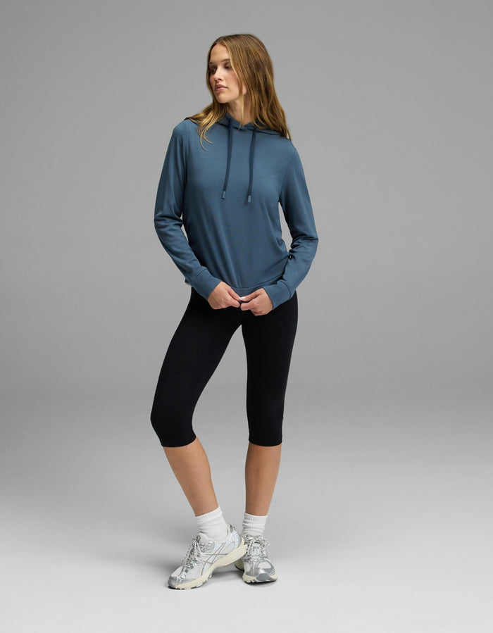 A woman stands on a gray background wearing the Cozy Earth Women’s Brushed Bamboo Hoodie, paired with black capri leggings, white socks, and white sneakers. She looks to the side with one hand in her hoodie pocket.|Color:Dawn