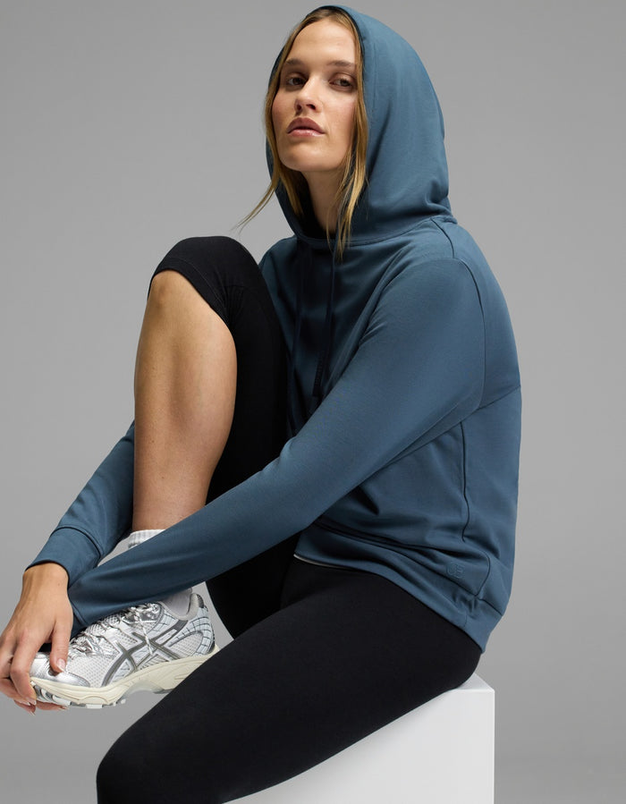 A woman wears the Cozy Earth Women’s Brushed Bamboo Hoodie, paired with black leggings and white sneakers, as she sits confidently on a white block against a gray background.|Color:Dawn