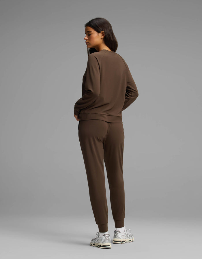 A woman models the Cozy Earth Women's Brushed Bamboo Jogger Set, featuring a brown long-sleeve top and matching jogger pants. She faces away and glances to her left, wearing white sneakers against a plain gray background.|Color:Espresso