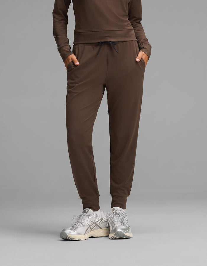 A person wearing the Cozy Earth Women's Brushed Bamboo Jogger Set in brown, paired with silver athletic sneakers, stands with hands in their pockets against a gray background.|Color:Espresso