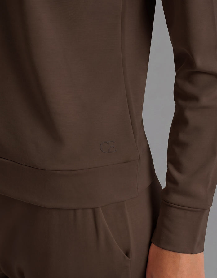A person wearing the Cozy Earth Women's Brushed Bamboo Jogger Set in brown stands with one arm relaxed. The smooth fabric is complemented by a small embossed logo near the hem of the long-sleeve top.|Color:Espresso