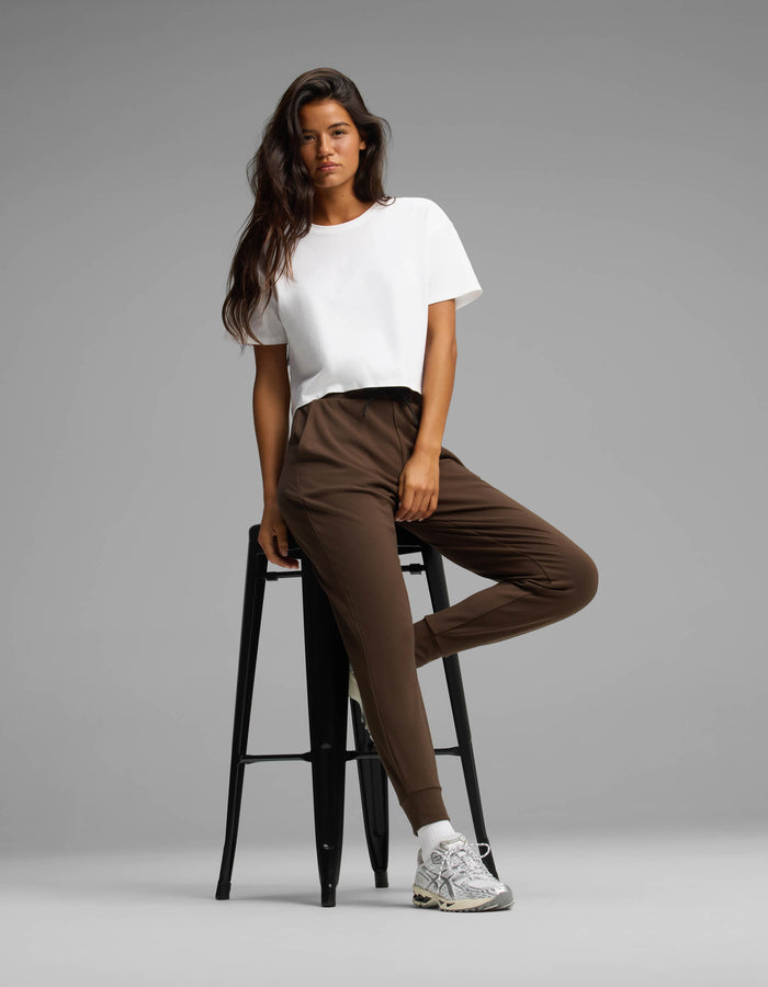 A woman models the Cozy Earth Women's Brushed Bamboo Joggers in brown, paired with a white t-shirt and white sneakers. She stands with one leg crossed, leaning on a tall black stool against a plain gray background.|Color:Espresso