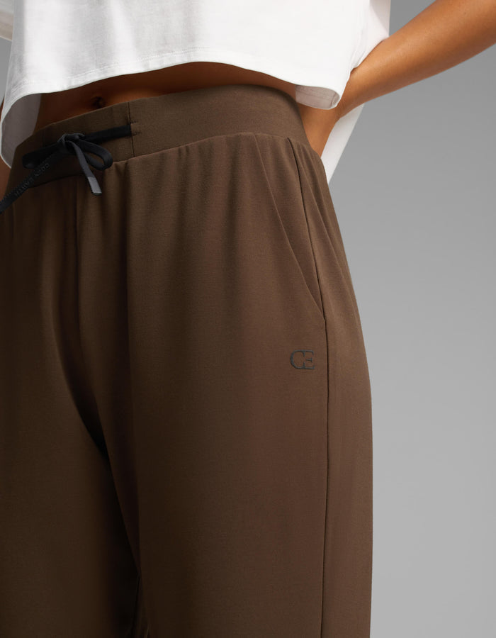 A person stands with one hand in their pocket, wearing Cozy Earth Women's Brushed Bamboo Joggers in brown paired with a white cropped shirt. The joggers feature a small logo near the pocket.|Color:Espresso