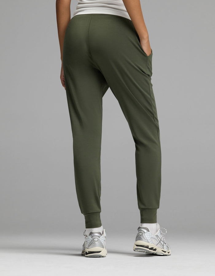 A model is shown from the back against a plain gray background, wearing Cozy Earth’s Women’s Brushed Bamboo Jogger in olive green paired with a white top and white sneakers.|Color:Olive