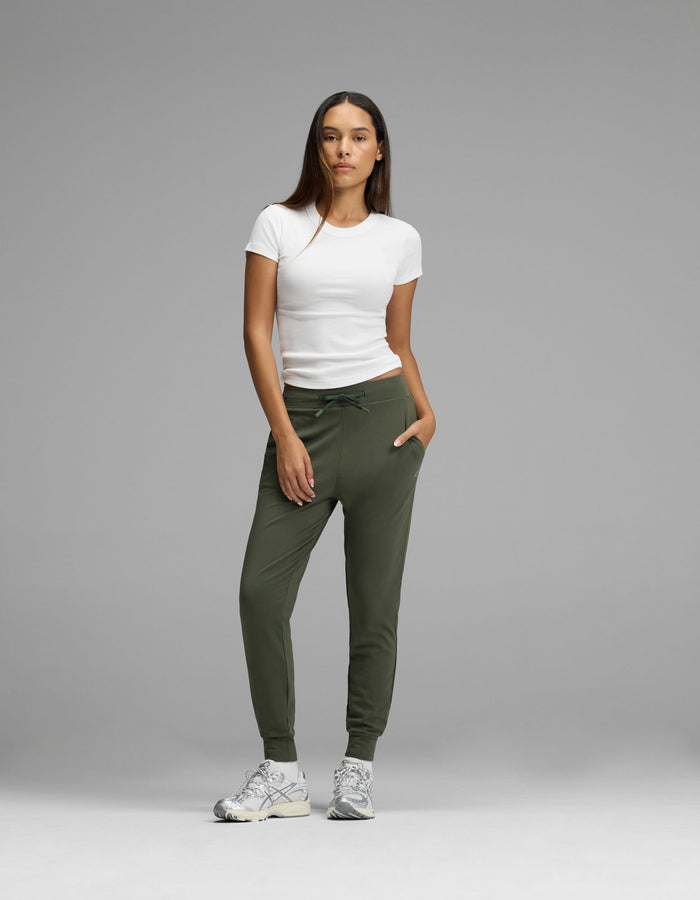A woman stands against a gray background wearing a white t-shirt, gray sneakers, and Cozy Earth’s Women’s Brushed Bamboo Jogger in olive green, with her hands in her pockets.|Color:Olive