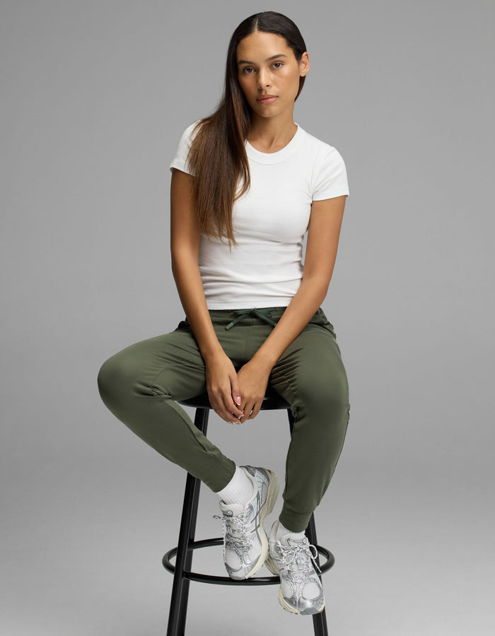 A woman with long brown hair sits on a black stool, wearing the Cozy Earth Women’s Brushed Bamboo Jogger in olive green, paired with a white t-shirt, white socks, and silver sneakers. She looks at the camera with a neutral expression against a plain gray background.|Color:Olive