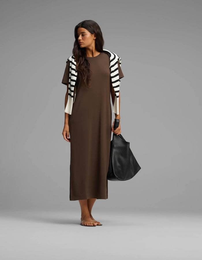 A woman stands barefoot against a plain background, wearing Cozy Earth’s Women's Brushed Bamboo Midi Dress in brown, with a black-and-white striped sweater over her shoulders and holding a large black bag.|Color:Espresso