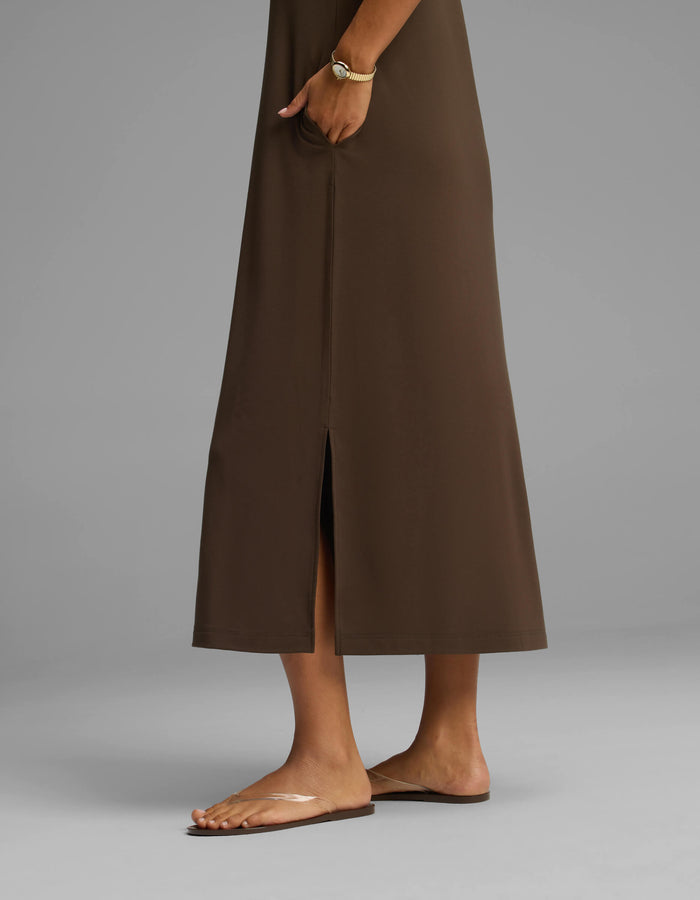 The lower half of a person is shown wearing the Cozy Earth Women's Brushed Bamboo Midi Dress in brown, featuring a center slit, paired with a bracelet and clear flat sandals, standing against a plain gray background.|Color:Espresso