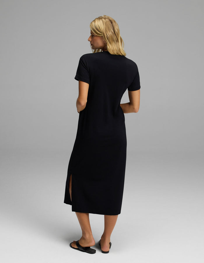 A woman with blonde hair stands facing away in a plain, light-colored studio, wearing Cozy Earth’s Women’s Brushed Bamboo Midi Dress—a black, short-sleeve style with a side slit—paired with black flats.|Color:Black