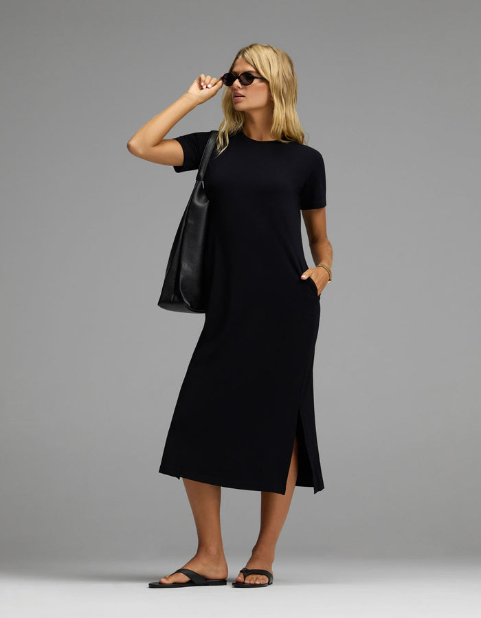 A woman stands confidently against a plain gray background, wearing the Cozy Earth Women’s Brushed Bamboo Midi Dress in black with short sleeves and side slits. She completes her look with black sandals, sunglasses, and a large black tote bag.|Color:Black