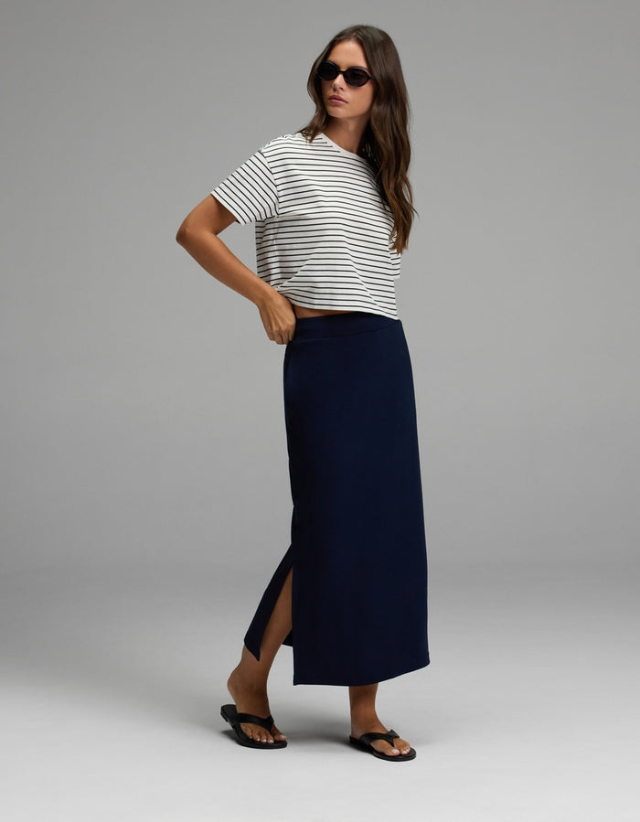 A woman wearing sunglasses, a white and black striped t-shirt, black sandals, and the Cozy Earth Women’s Brushed Bamboo Midi Skirt in navy blue with a side slit, standing against a plain gray background.|Color:Navy