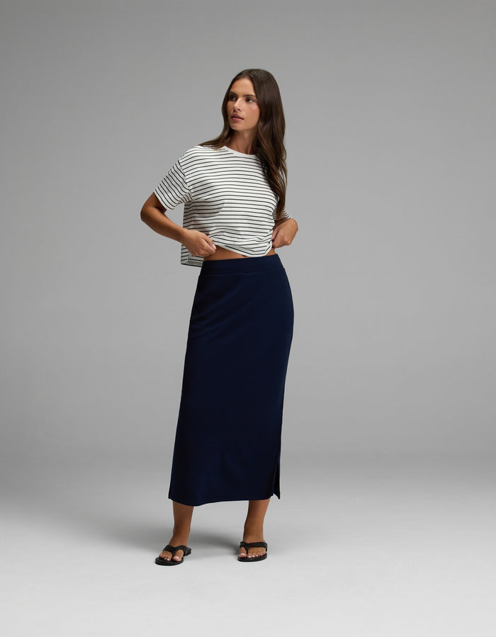 A woman stands in a studio wearing a black and white striped t-shirt, the Cozy Earth Women’s Brushed Bamboo Midi Skirt in navy with a side slit, and black sandals. She looks to the side with one hand adjusting her shirt.|Color:Navy