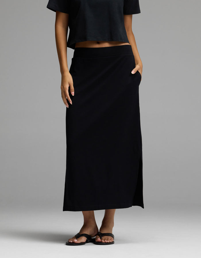 A woman wearing Cozy Earth’s Women’s Brushed Bamboo Midi Skirt in black, paired with a black crop top and black sandals, stands with one hand in her skirt pocket against a plain gray background.|Color:Black
