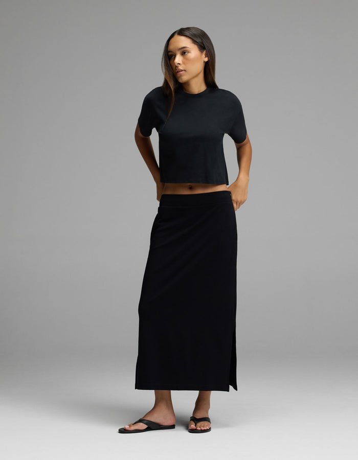 A woman stands in a studio wearing the Cozy Earth Women’s Brushed Bamboo Midi Skirt in black, paired with a black short-sleeve crop top and black sandals against a neutral background.|Color:Black