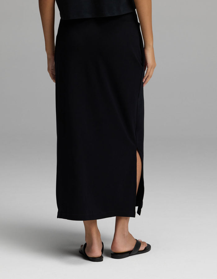 A person is shown from the back, wearing Cozy Earth’s Women’s Brushed Bamboo Midi Skirt in black—with a side slit—paired with black sandals, standing against a light gray background. Their upper body is partially visible.|Color:Black
