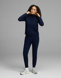 A woman wearing the Cozy Earth Women’s Brushed Bamboo Mock Neck & Jogger Set in navy stands against a gray background, adjusting the drawstrings of her hoodie and pairing the outfit with white sneakers.|Color:Navy