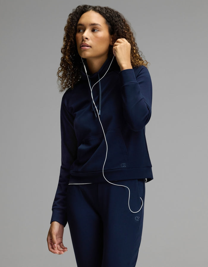 A woman with curly hair, dressed in the Cozy Earth Women’s Brushed Bamboo Mock Neck & Jogger Set in navy, listens to wired earphones—holding one earbud and gazing to the side against a plain gray background.|Color:Navy