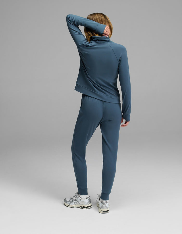 A person wears the Cozy Earth Women’s Brushed Bamboo Mock Neck & Jogger Set, paired with white sneakers, standing with one arm bent and covering part of their face, against a plain gray background.|Color:Dawn