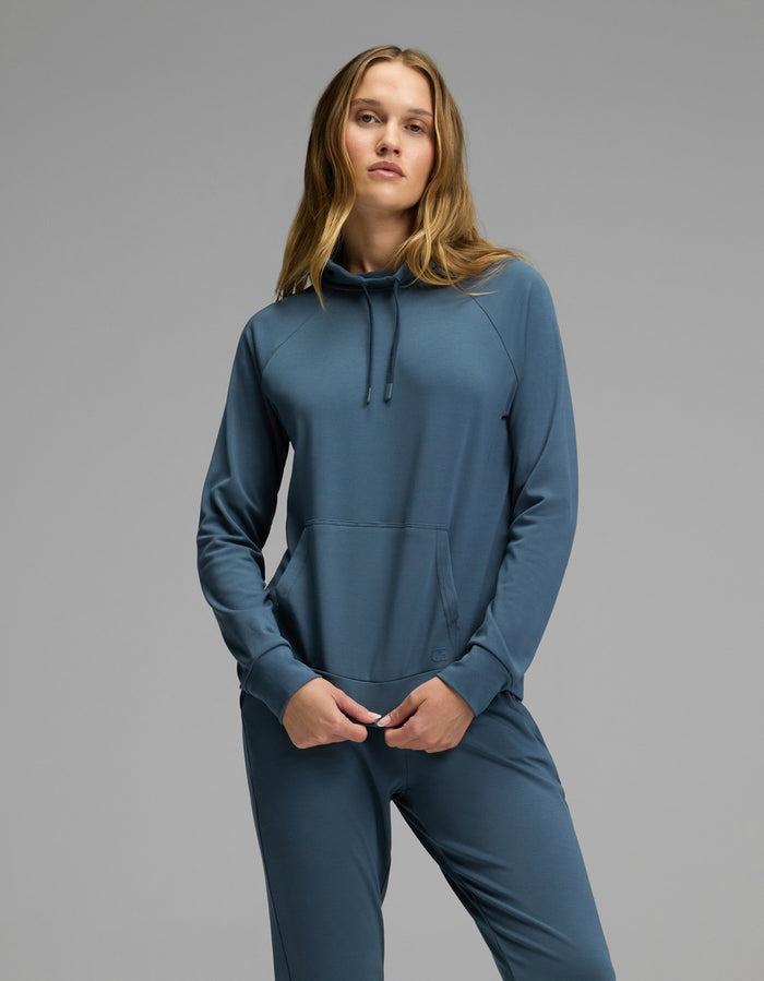 A woman models the Cozy Earth Women’s Brushed Bamboo Mock Neck in teal, standing confidently with her hands in the pockets against a plain gray background.|Color:Dawn