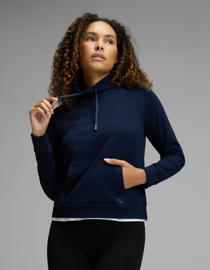 A woman with curly hair wears the Cozy Earth Women’s Brushed Bamboo Mock Neck in navy, paired with black leggings. She gazes upward and slightly to the side, holding a drawstring with one hand while her other hand rests in the front pocket, set against a neutral gray background.|Color:Navy
