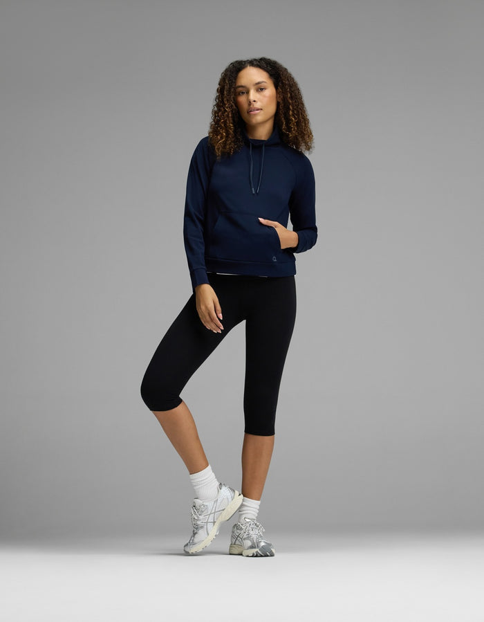 A woman with curly hair stands against a gray background with one leg slightly bent, wearing the Cozy Earth Women’s Brushed Bamboo Mock Neck in navy, paired with black capri leggings, white socks, and sneakers.|Color:Navy