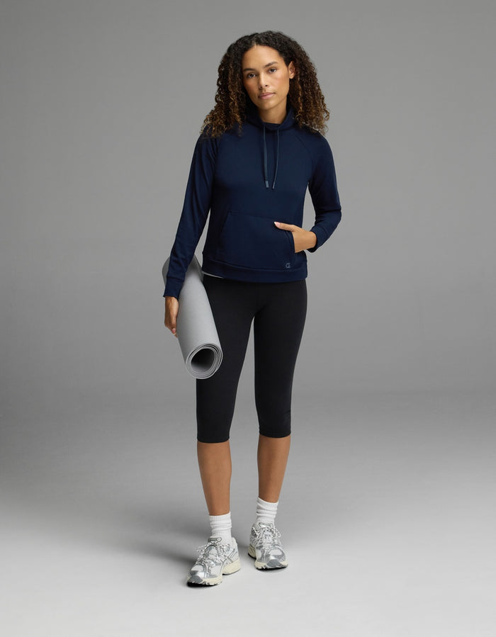 Wearing the Cozy Earth Women’s Brushed Bamboo Mock Neck in navy with black leggings, a person stands confidently against a gray background, holding a rolled-up yoga mat in one hand. They complete the look with white sneakers and have curly hair, gazing self-assuredly at the camera.|Color:Navy