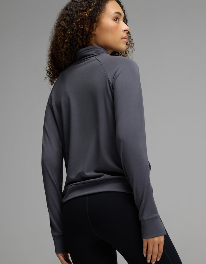 A person with curly hair wearing the Cozy Earth Women’s Brushed Bamboo Mock Neck in dark gray and black leggings stands facing away, looking over their shoulder against a plain gray background.|Color:Graphite