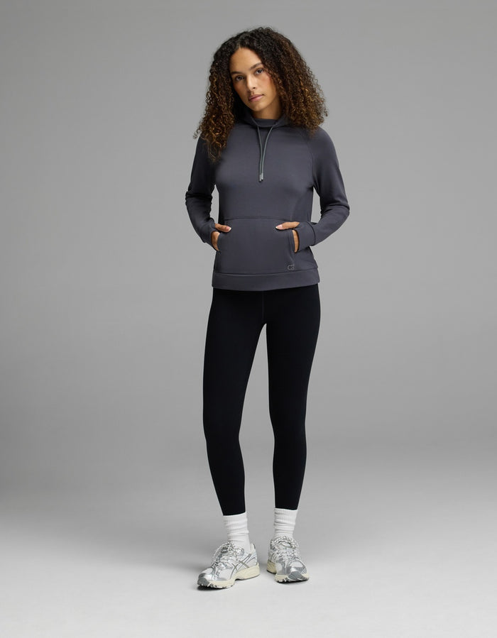 A person stands against a plain gray background, wearing the Cozy Earth Women’s Brushed Bamboo Mock Neck with their hands in the sweatshirt pockets, paired with black leggings, white socks, and white sneakers. Their curly hair falls loosely around their shoulders.|Color:Graphite