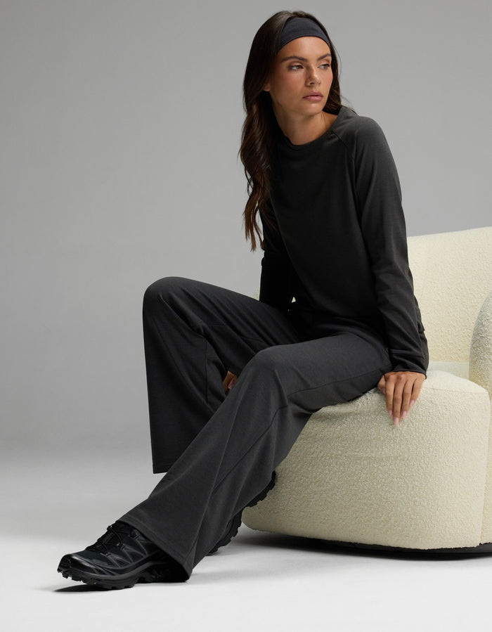 A woman models the Cozy Earth Women’s Brushed Bamboo Wide Leg Pull On Pant & Pullover Crew Set in a dark hue, paired with black shoes and a matching headband. She sits on a cream-colored chair, gazing to the side against a plain gray background.|Color:Charcoal