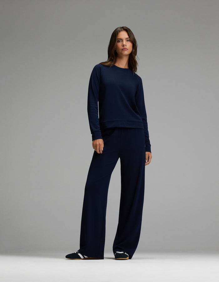 A woman stands against a gray background wearing the Cozy Earth Women’s Brushed Bamboo Wide Leg Pull On Pant & Pullover Crew Set in navy, paired with black slide sandals.|Color:Navy