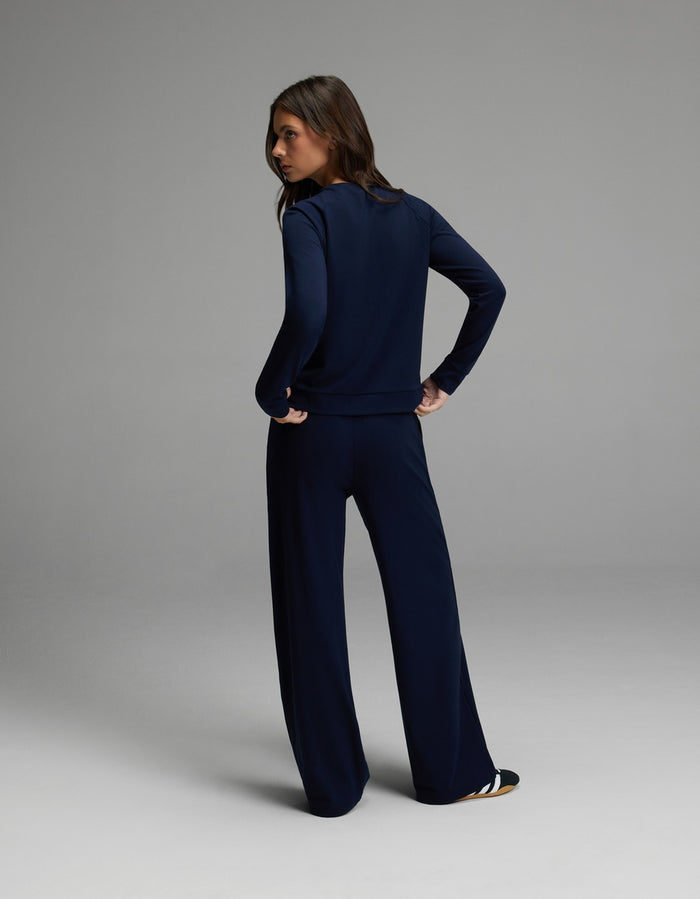 A woman stands facing away with her hands on her hips, wearing the Cozy Earth Women’s Brushed Bamboo Wide Leg Pull On Pant & Pullover Crew Set in navy. The background is a simple, light gray studio setting.|Color:Navy