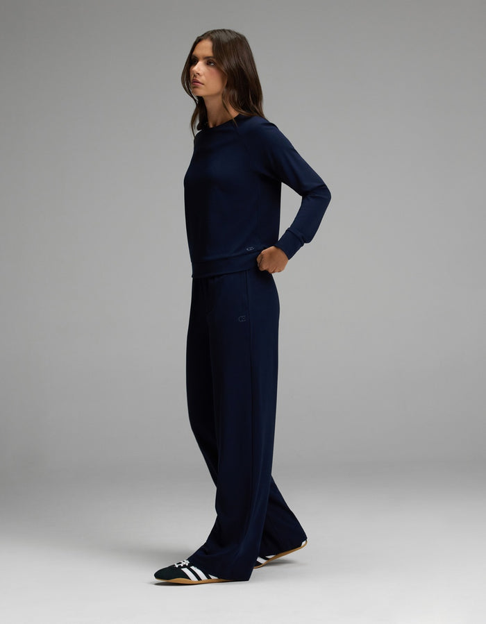 A woman stands sideways against a plain gray background, wearing the Cozy Earth Women’s Brushed Bamboo Wide Leg Pull On Pant & Pullover Crew Set in dark blue, paired with black and white slide sandals.|Color:Navy
