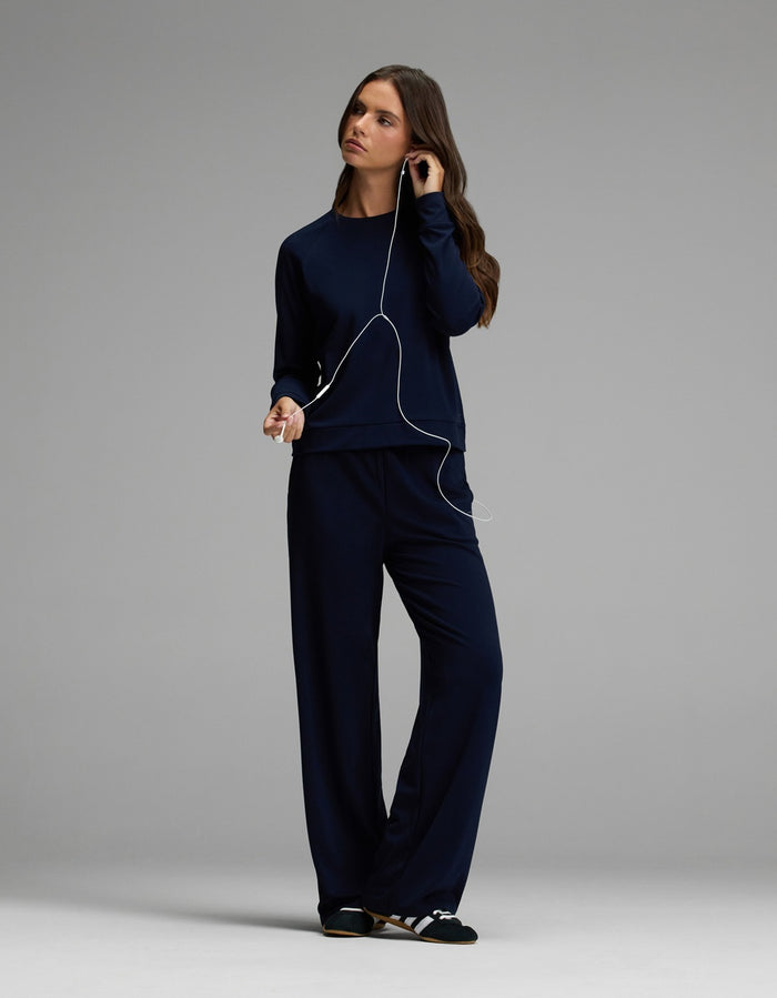 A woman wearing the Cozy Earth Women’s Brushed Bamboo Wide Leg Pull On Pant & Pullover Crew Set in navy and striped slippers stands against a gray background, listening to music with wired earphones.|Color:Navy
