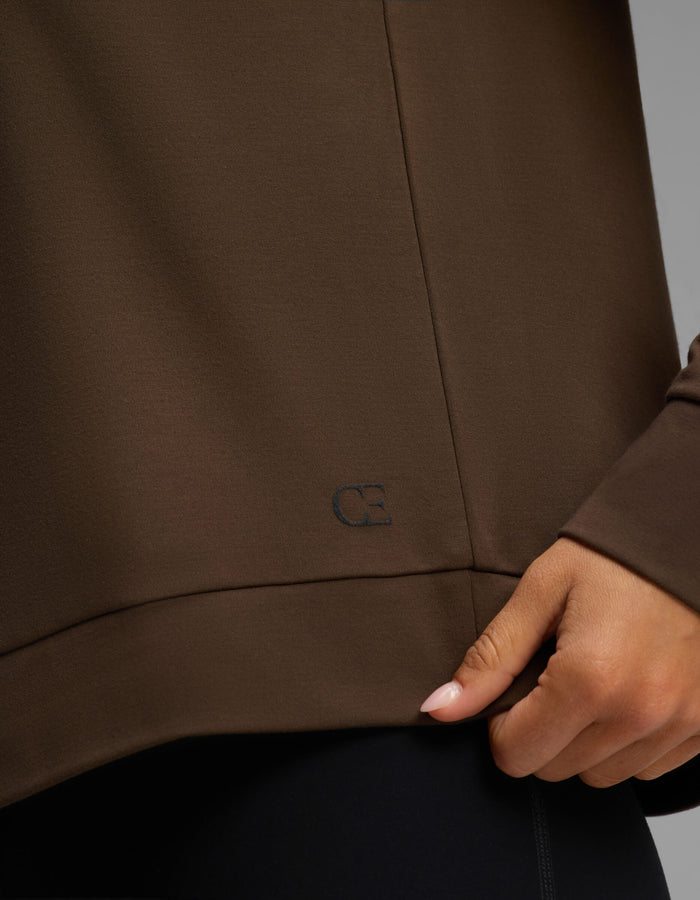A person wearing the Cozy Earth Women's Brushed Bamboo Pullover Crew in brown gently pinches the hem with one hand, revealing a small, subtle embroidered "CB" logo near the bottom edge.|Color:Espresso