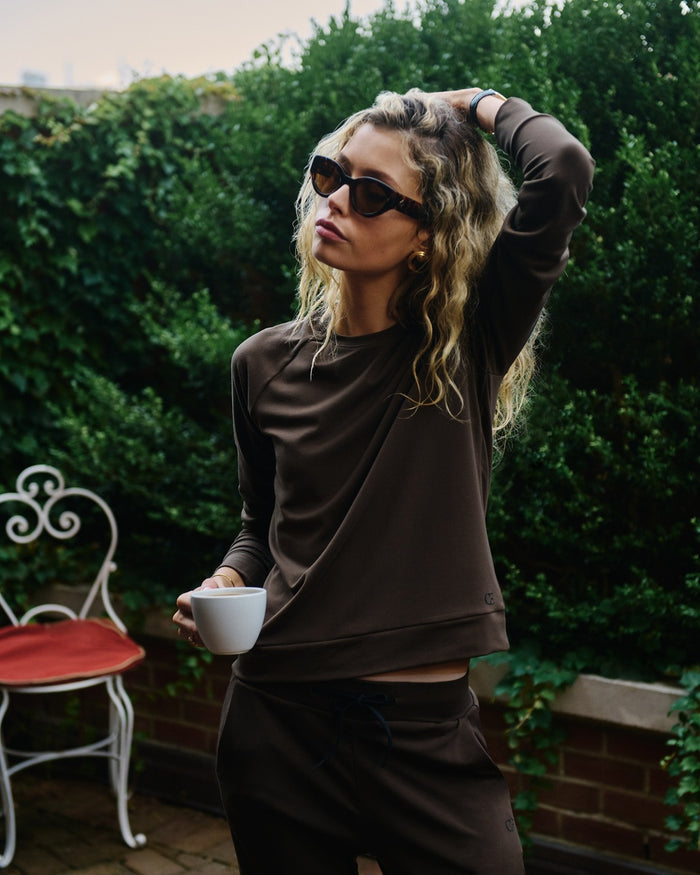 A woman wearing a brown Cozy Earth Brushed Bamboo Pullover and Jogger set, styled with black sunglasses and holding a white mug, standing outdoors in front of a green hedge. |Color:Espresso