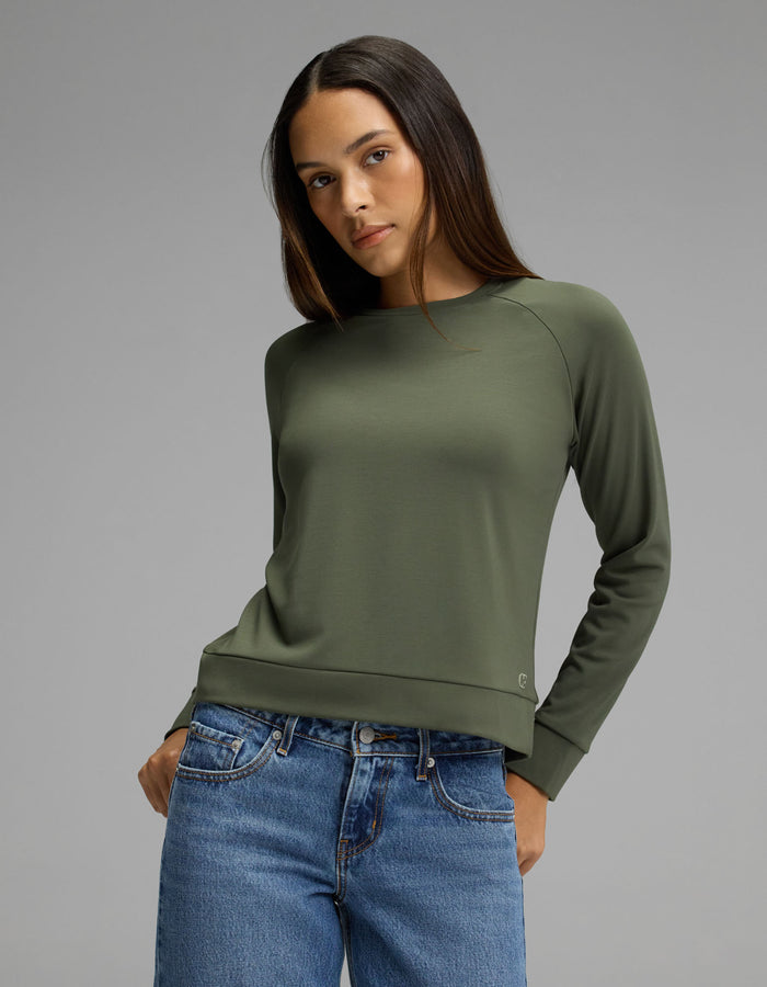 A woman models the Cozy Earth Women’s Brushed Bamboo Pullover Crew in olive, paired with blue jeans, as she stands against a plain gray background and looks slightly to the side with one hand in her pocket.|Color:Olive