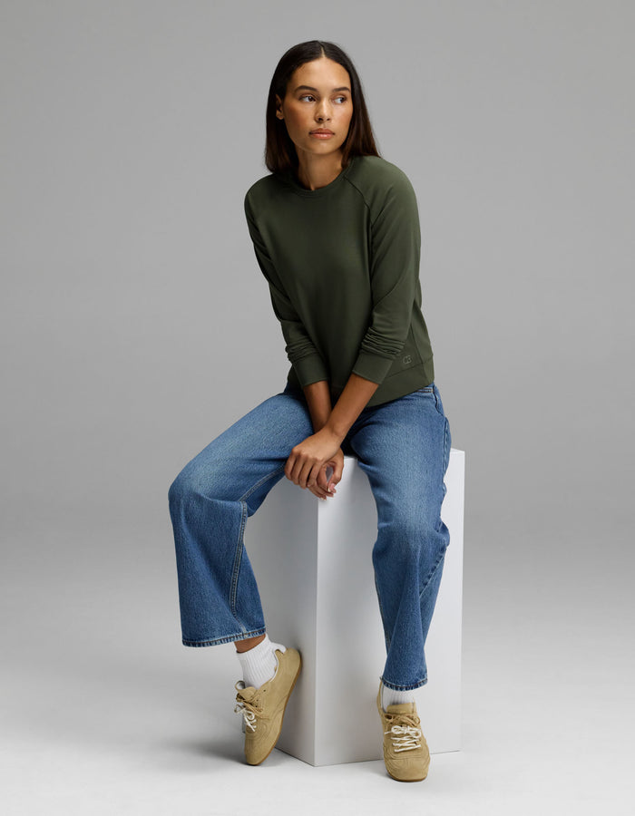A woman wears the Cozy Earth Women’s Brushed Bamboo Pullover Crew in green and blue jeans as she sits on a white block against a plain gray background, looking slightly to her left. She pairs her outfit with beige sneakers and white socks.|Color:Olive