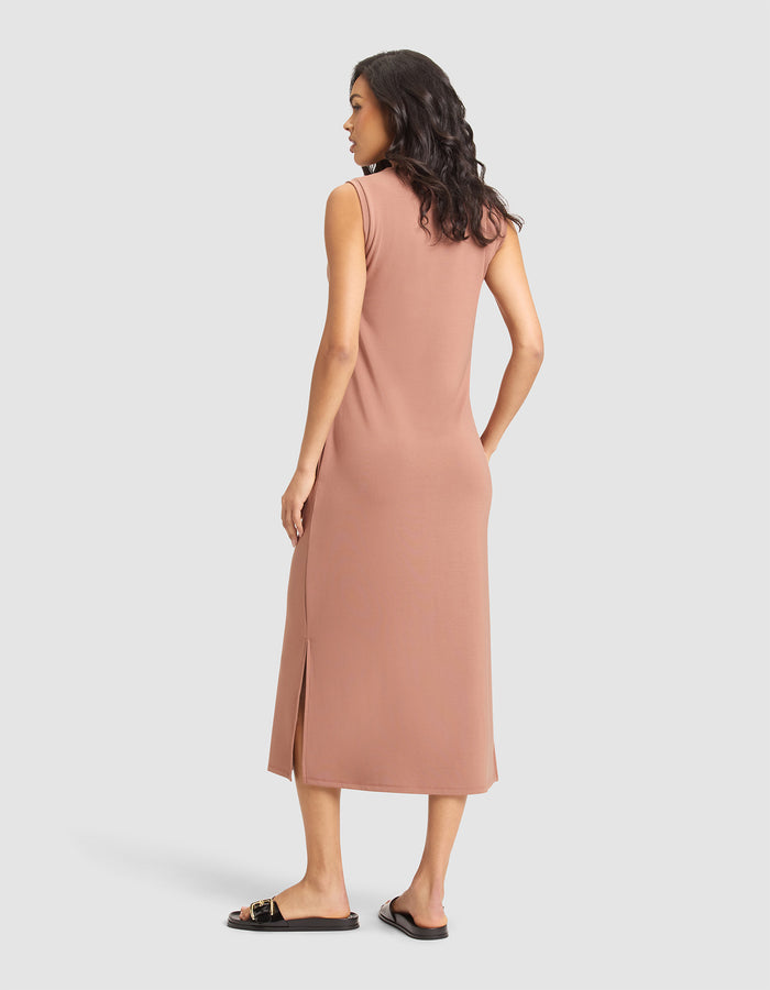 A woman with wavy dark hair, wearing black flat sandals and the Cozy Earth Women's Brushed Bamboo Sleeveless Midi Dress in rose, stands facing away against a plain light gray background. The knee-length dress features a side slit. |Color:Saddle