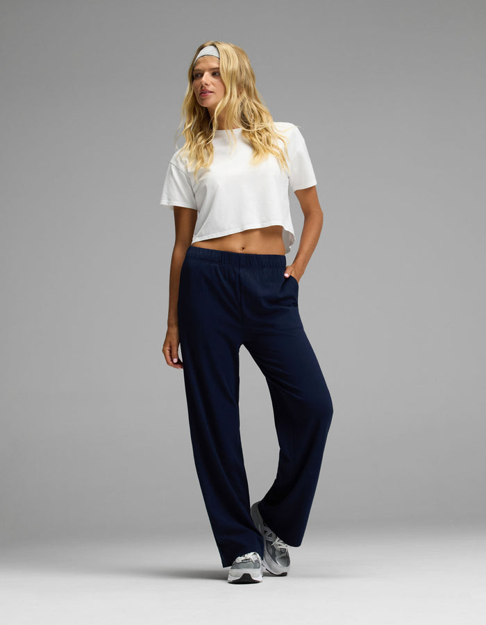 A woman stands against a plain gray background with one hand in her pocket, wearing Cozy Earth’s Women’s Brushed Bamboo Wide Leg Pull-On Pant in navy blue, paired with a white cropped t-shirt, gray sneakers, and a white headband.|Color:Navy