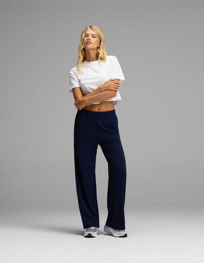 A person stands against a plain background, wearing a white cropped t-shirt, white sneakers, and the Cozy Earth Women’s Brushed Bamboo Wide Leg Pull-On Pant in dark blue. Their long blonde hair falls over their shoulders as they cross their arms and look off to the side.|Color:Navy