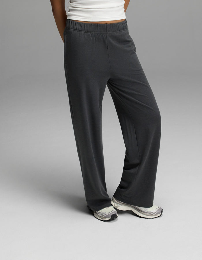 A woman wears Cozy Earth’s Women’s Brushed Bamboo Wide Leg Pull-On Pant in Charcoal, paired with white sneakers and a white top, standing with one leg crossed in front of the other against a plain gray background.|Color:Charcoal