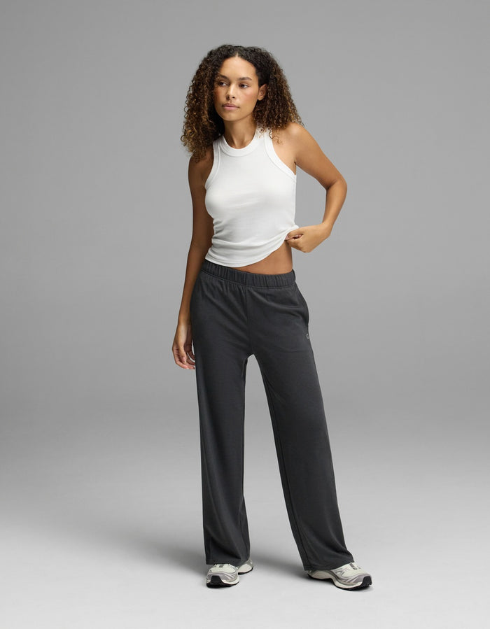 A woman with curly hair stands against a plain gray background, wearing a white sleeveless top and the Cozy Earth Women’s Brushed Bamboo Wide Leg Pull-On Pant in dark gray, paired with sneakers. She holds up one side of her shirt slightly with one hand.|Color:Charcoal