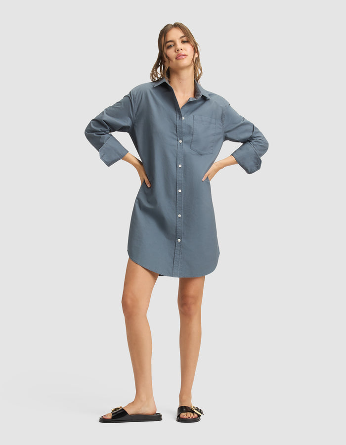 A woman wears the Cozy Earth Women's Camden Dress, a loose-fitting blue button-down shirt-dress, paired with black slide sandals with gold accents. Her long hair is pulled back and she stands against a plain light background. |Color:Dawn