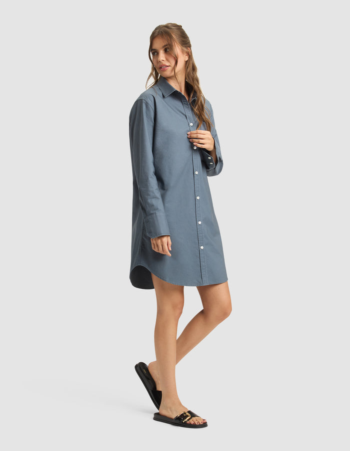 A woman poses in a relaxed way, wearing Cozy Earth's Women's Camden Dress—a loose-fitting, long-sleeve blue shirt dress—paired with black sandals featuring gold accents against a plain light background. |Color:Dawn