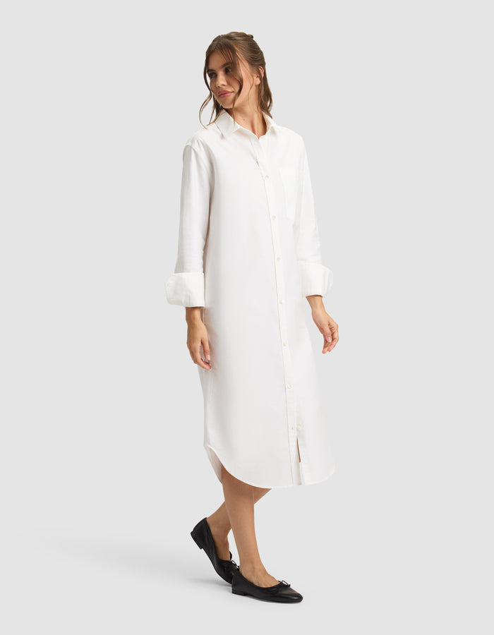 A woman models the Cozy Earth Women's Camden Midi Dress—a long white button-down shirt dress with rolled-up sleeves—paired with black flats, standing against a plain light gray backdrop. |Color:White