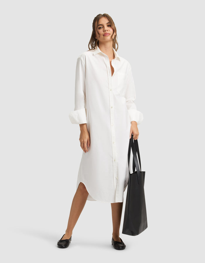 A woman smiles in a Cozy Earth Women's Camden Midi Dress, holding a large black tote bag. Her long brown hair is tied back and she wears black flats against a plain light gray background. |Color:White